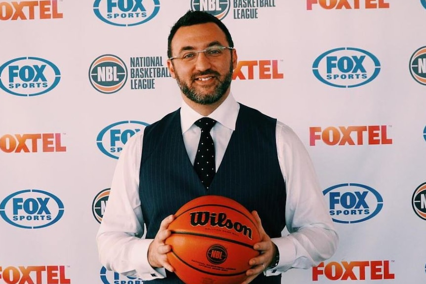 NBL CEO Jeremy Loeliger stands holding a basketball in front of a media wall.