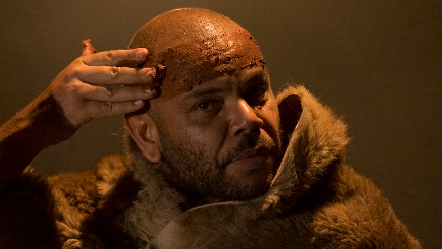 An Indigenous Australian man wearing a fur smears coloured clay across his forehead, beneath a yellow light.