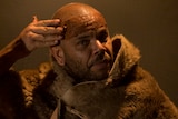 An Indigenous Australian man wearing a fur smears coloured clay across his forehead, beneath a yellow light.