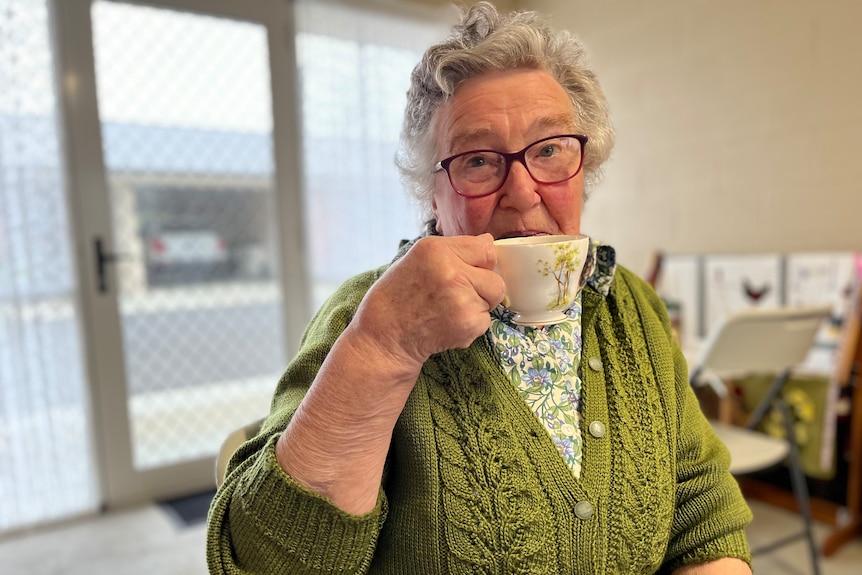 Lady with white hair wearing a green cardigan, holding a cup of tea 