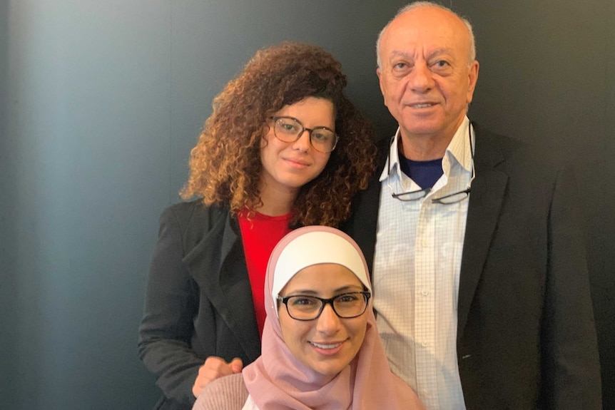 A young woman in a pink hijab sits in front of another young woman with curly hair and an older man.