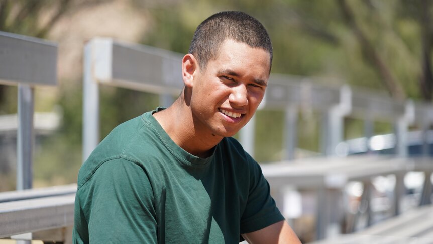 A young man wearing a green prison shirt squints into the sun and smiles at the camera while seated on a granstand.