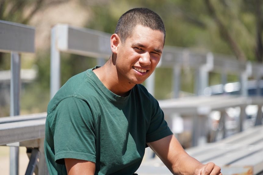 A young man wearing a green prison shirt squints into the sun and smiles at the camera while seated on a granstand.