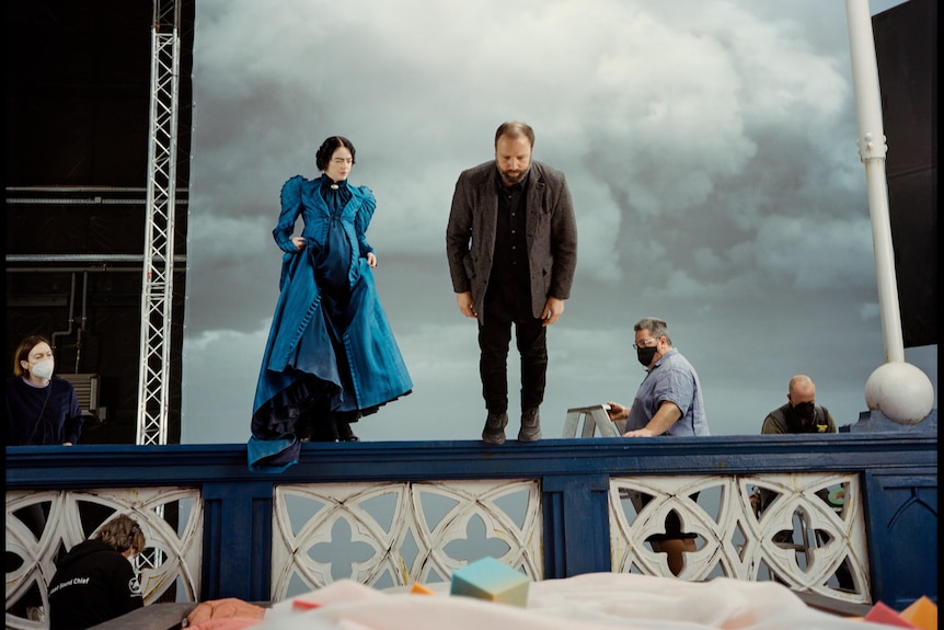 Emma, dressed in a blue gown, looks on as Yorgos stands on the ledge of a fake bridge and looks down.