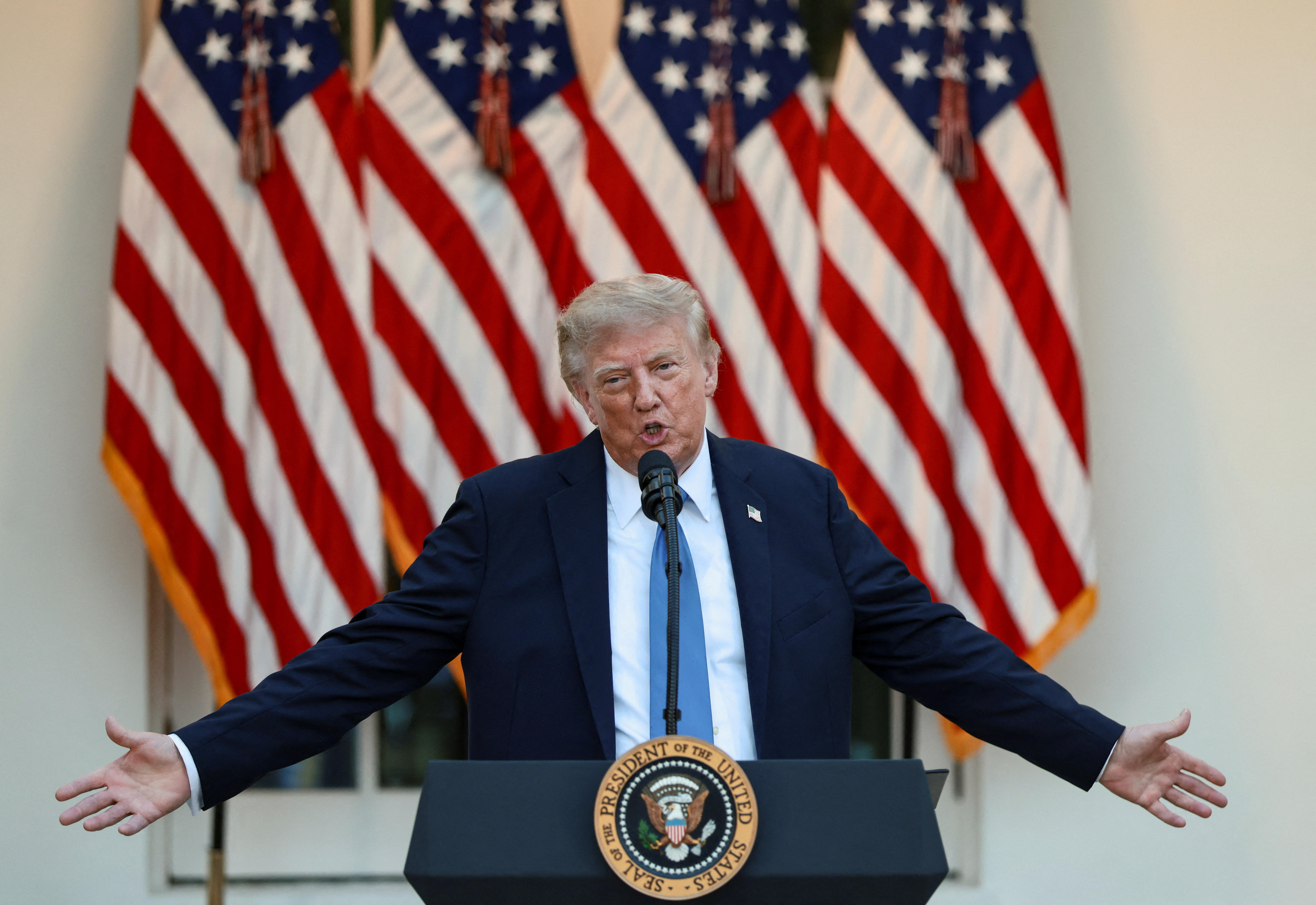Donald Trump in a black suit, white shirt and blue tie speaking at a lectern with his arms outstretched in front of US flags.