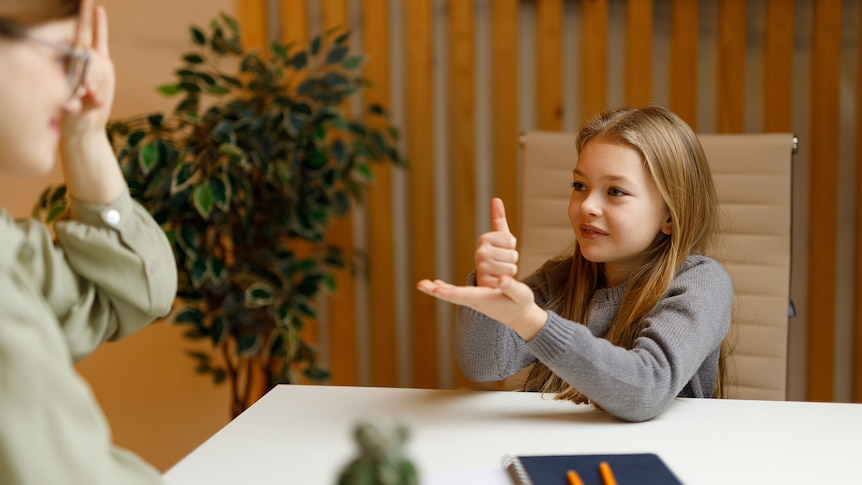 A young girl in a classroom communicates with a teacher using sign language.
