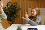 A young girl in a classroom communicates with a teacher using sign language.
