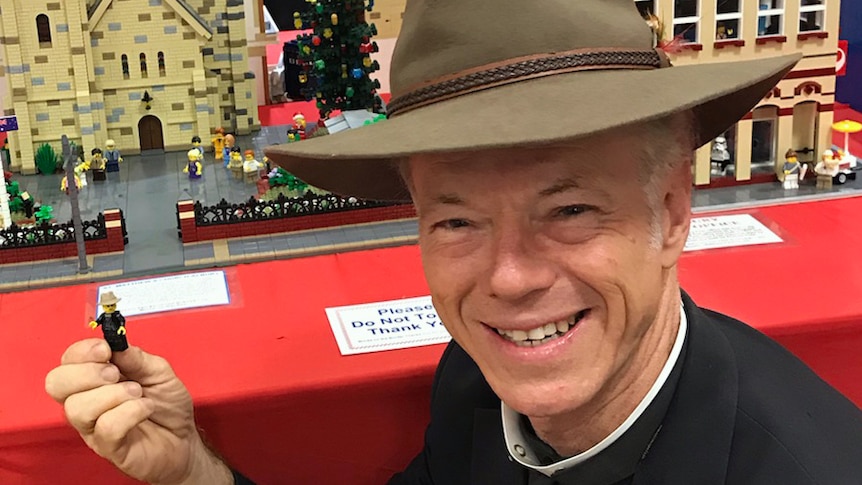 Priest in front of photo, holds a Lego version of himself with a Lego church model in the background.