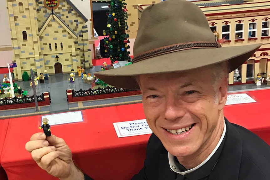Priest in front of photo, holds a Lego version of himself with a Lego church model in the background.