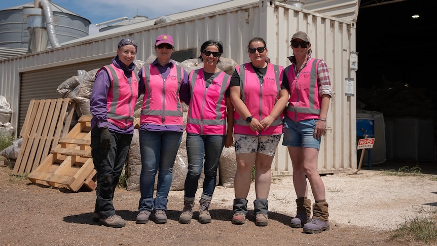Five women posing and smiling at the camera in front of a shed, smiling at the camera.