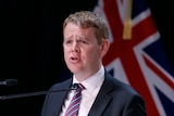 New Zealand's COVID-19 Response Minister Chris Hipkins speaks in front of a New Zealand flag.