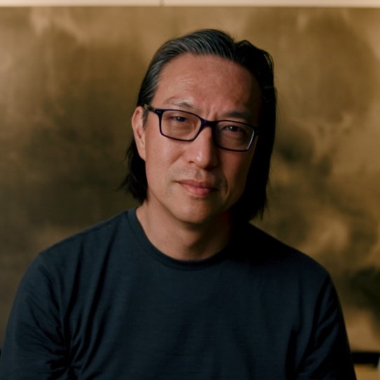 portrait of middle aged man with asian heritage wearing glasses sitting in front of large gold and black abstract painting