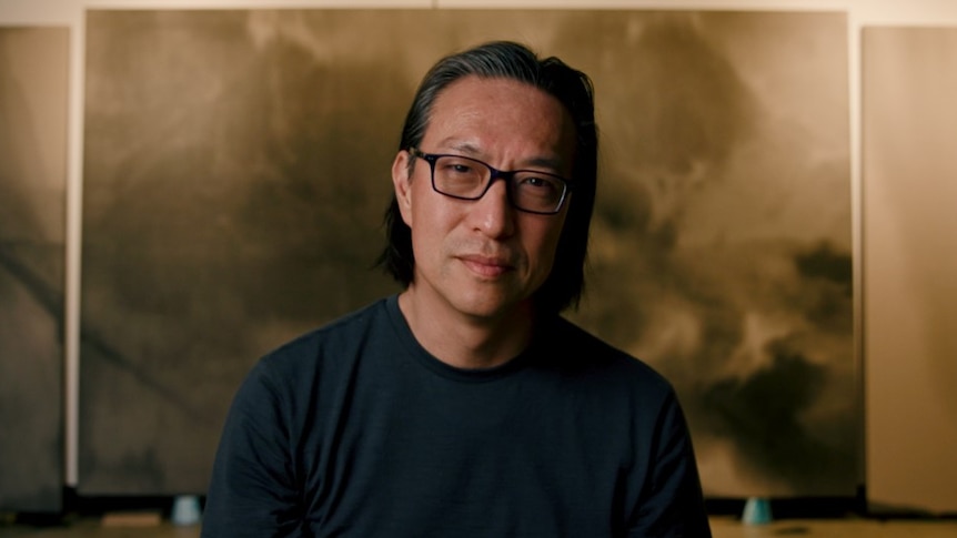 portrait of middle aged man with asian heritage wearing glasses sitting in front of large gold and black abstract painting