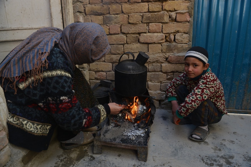 A woman and a little boy squat next to an outdoor fire with a pot hanging over it.