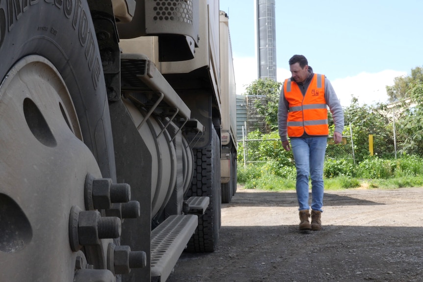 A man in a hi-vis orange vest and blue jeans inspects a large truck.