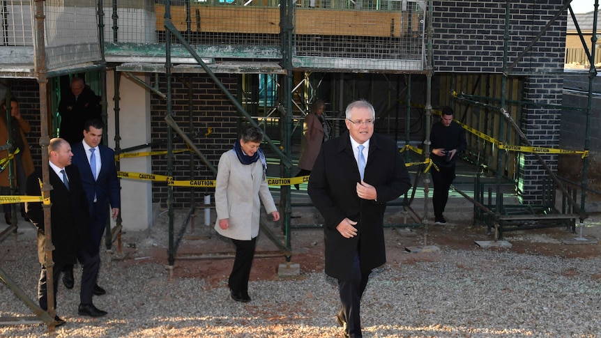 Scott Morrison, wearing a long winter jacket, walks out of a construction site followed by two men and a woman.