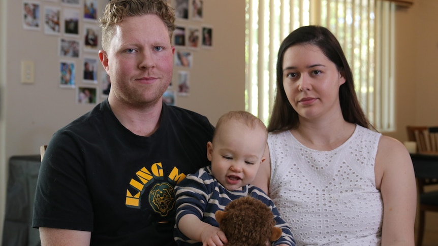 A man and a woman holding their baby son, in a living room, look into the camera with neutral expressions.
