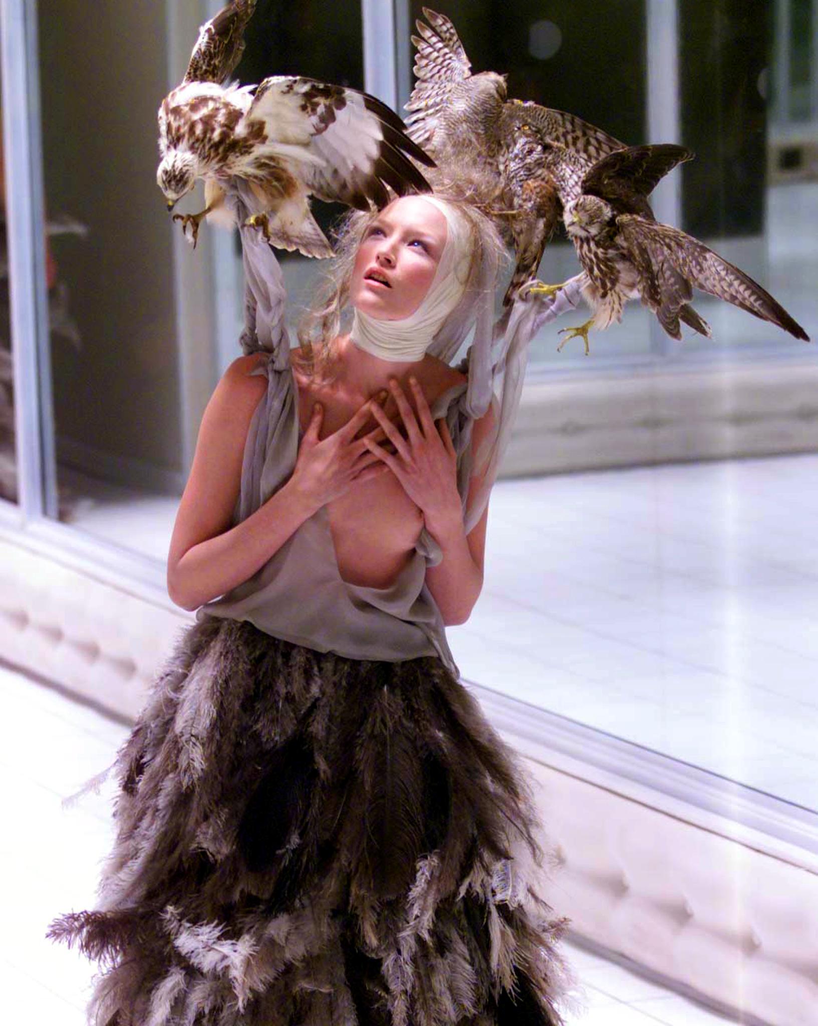 A young white model with her head wrapped in white fabric, with taxidermy birds on her shoulders.
