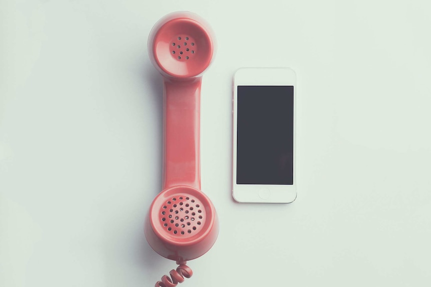 The pink receiver of an old phone alongside an iPhone on a white table..