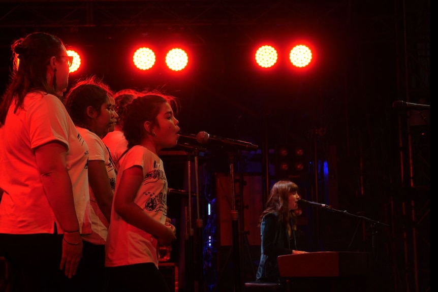 Girls in a group wearing white shirts singing in front of a microphone, with Missy Higgins wearing black singing in the back