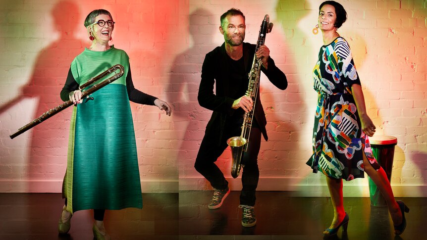 Three people against a white brick wall. One woman holds an alto flute, a man holds a bass clarinet.