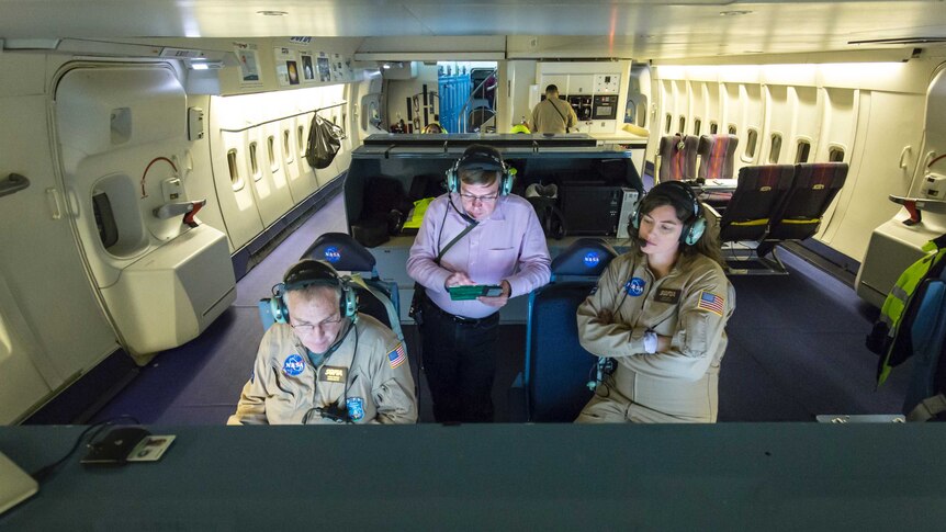 A man in a pink shirt and radio headset stands between two people in NASA jumpsuits in the cabin of an aeroplane.