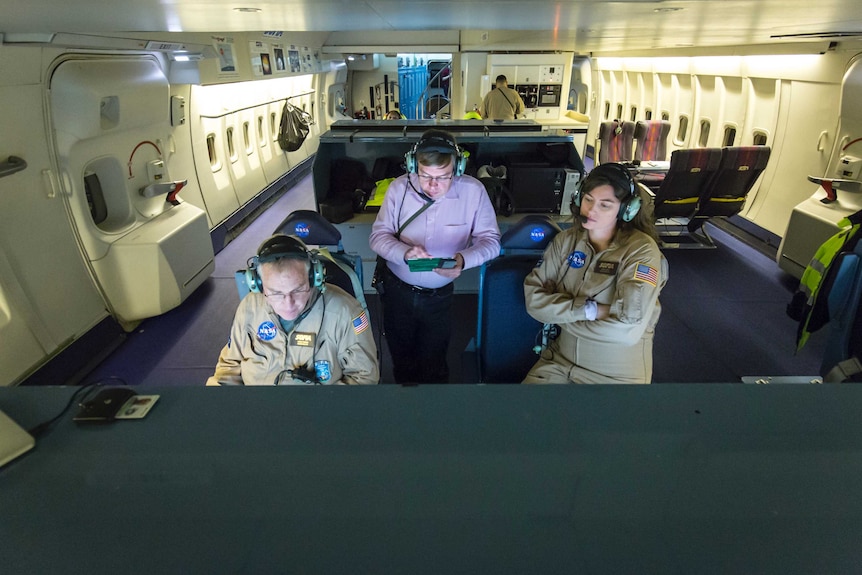 A man in a pink shirt and radio headset stands between two people in NASA jumpsuits in the cabin of an aeroplane.