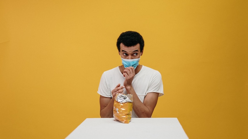 Against a yellow backdrop, you see a man sitting at a white table while clutching his light blue facemask.