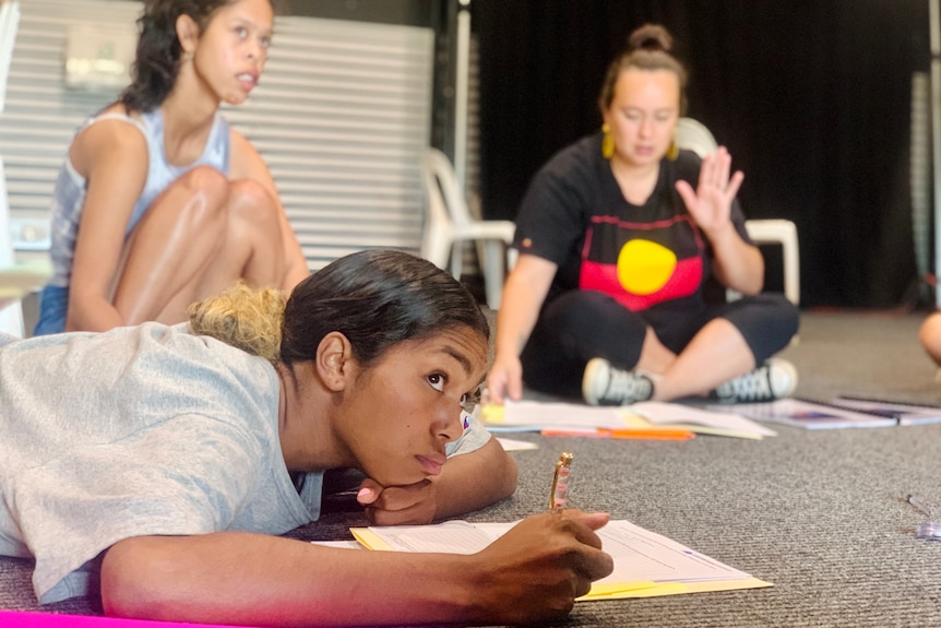 A teenager lays on the floor listening attentively with a workbook in front of her.