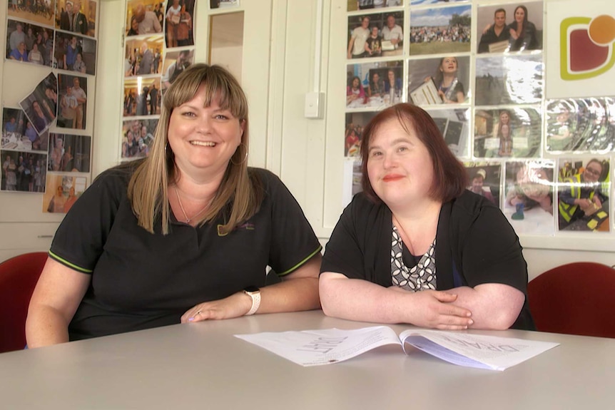 Two women sit at a desk; a sign behind reads 'ACT Down Syndrome Association'.