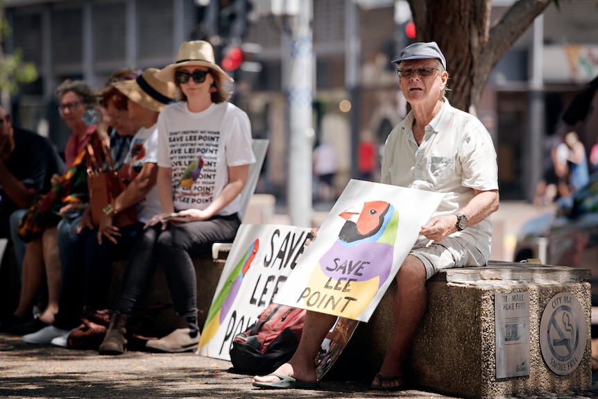 A man sitting on a bench, near several other people, and holding a sign with a painted picture of a finch. 