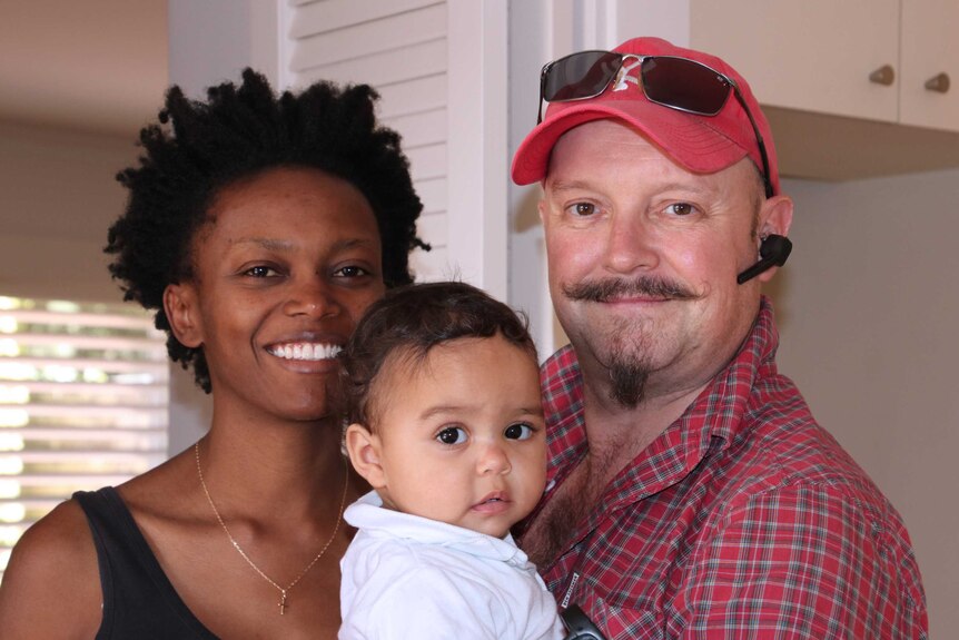 A mid-shot showing Perth couple Njeri and Shane posing for a photo in their house holding their 10-month-old daughter Amani.