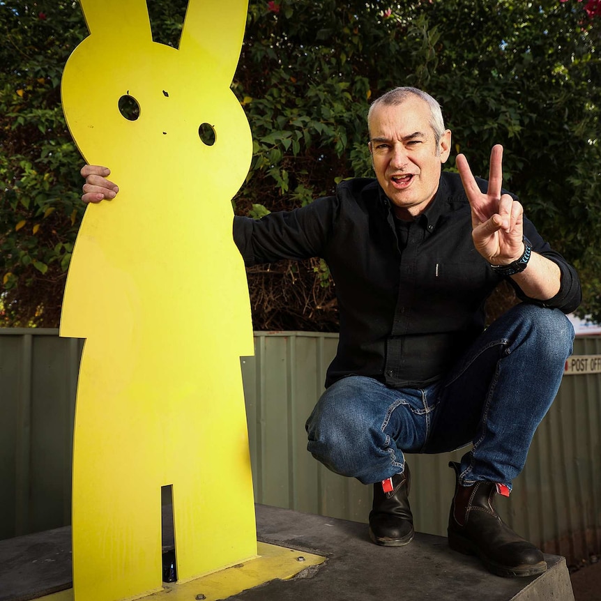 A man with short grey hair crouching next to a yellow silhouette of a rabbit, holding up two fingers to signify peace.