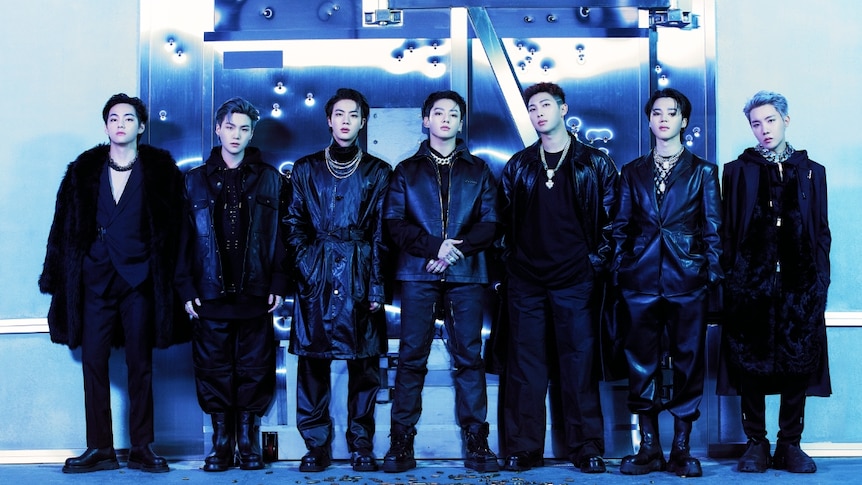 Seven South Korean boy band members standing in black outfits with blue lighting