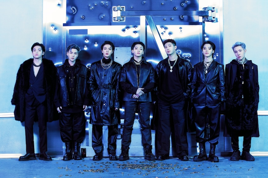 Seven South Korean boy band members standing in black outfits with blue lighting