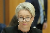 Bridget McKenzie, wearing glasses, sits at a table in a wood-panelled room with a folder open in front of her