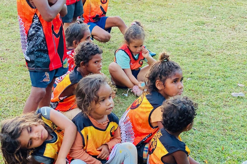 Indigenous children sitting on grass in orange,  yellow and black jersey.