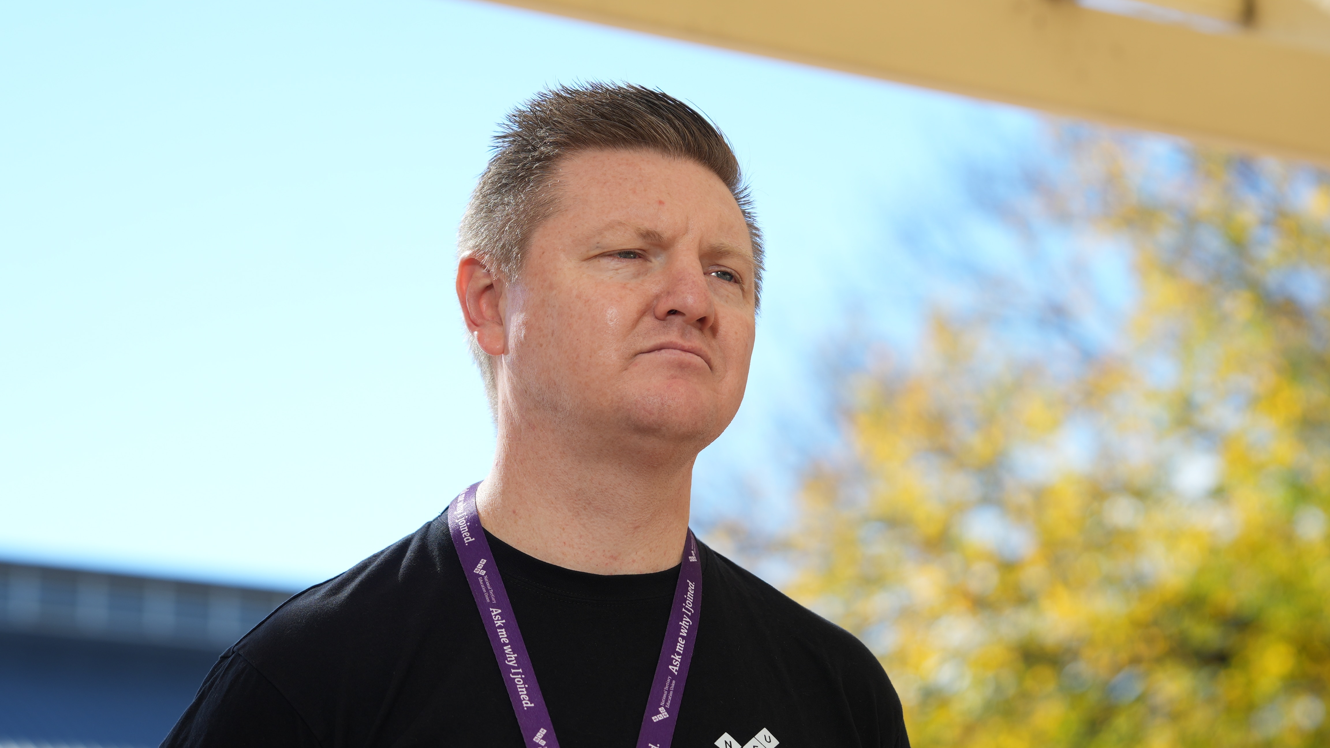 A man with short hair stands outdoors looking serious.