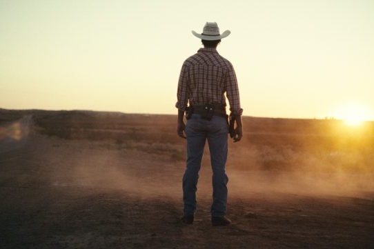 Man who looks like a cowboy stands with back to camera watching sunset. 