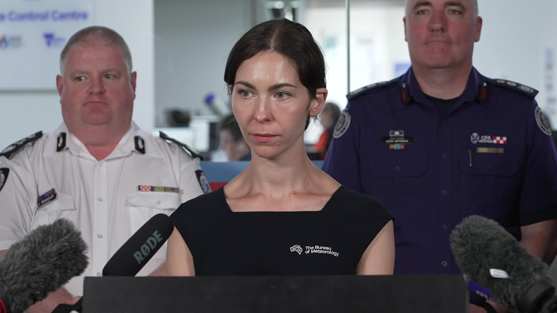 A woman with dark hair in a black dress stands behind microphones with two men standing behind her.