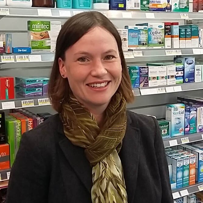 Katie Stott has short brown hair is wearing a dark green scarf and stands in front of shelves of medicine.