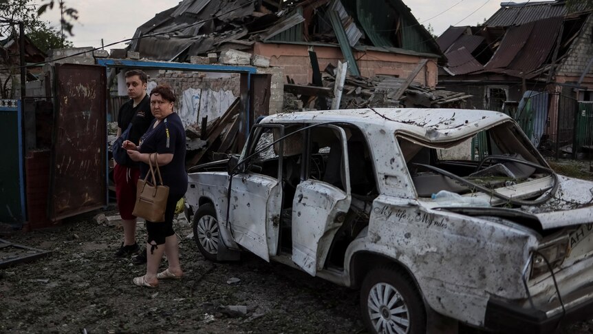 A man and a woman stand beside a destroyed car outside a shelled home.
