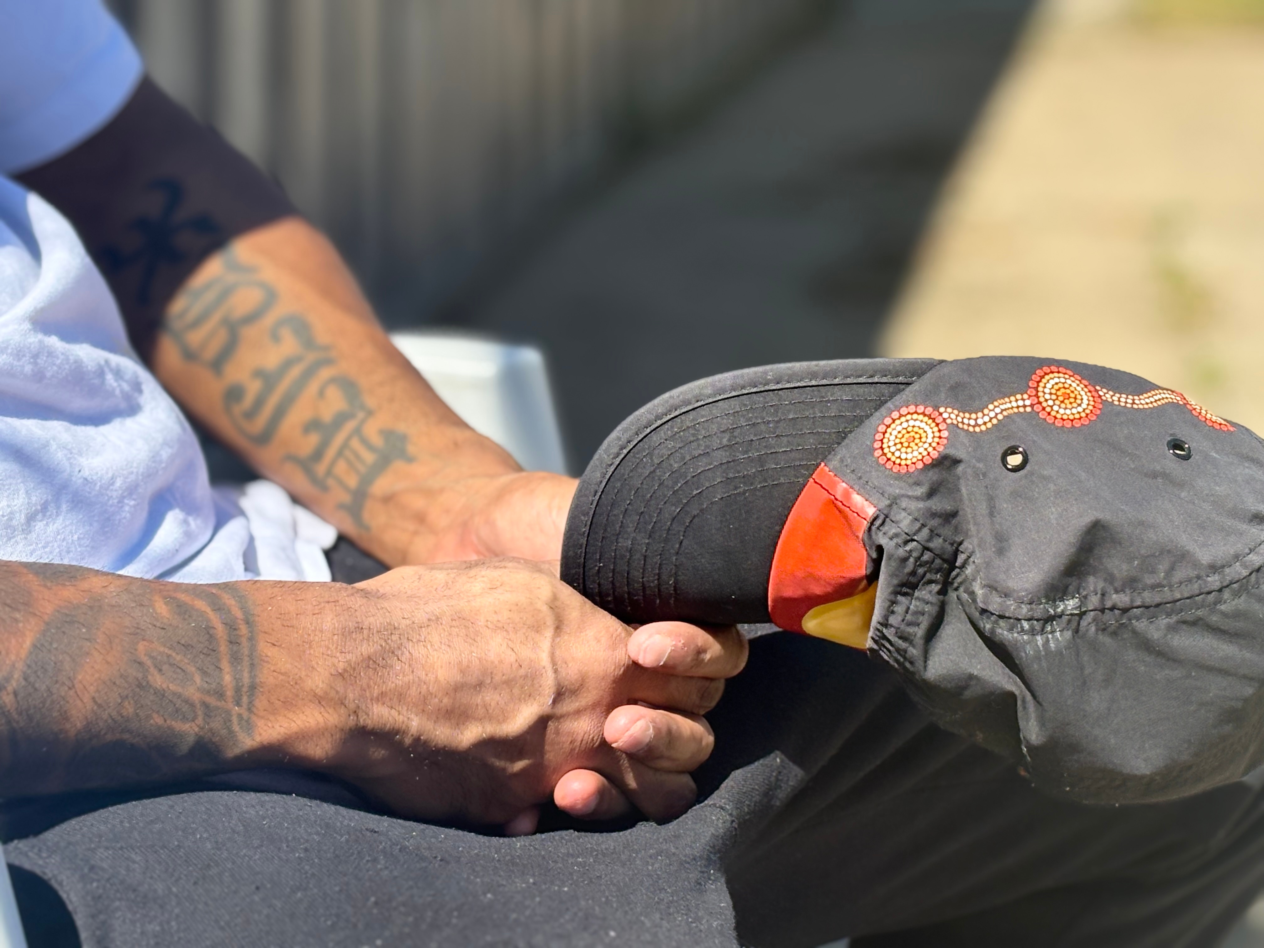 Uma foto em close das mãos de um homem segurando um chapéu com a bandeira aborígene. 