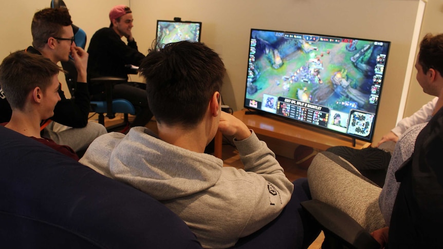 One man in a red cap, and four other men, looking at a screen where League of Legends is playing on-screen.