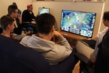 One man in a red cap, and four other men, looking at a screen where League of Legends is playing on-screen.