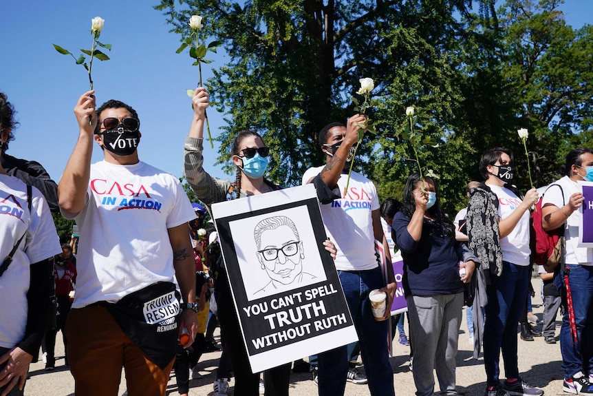 People hold white roses in the air, wearing masks and sunglasses. One has a sign reading 'You can't spell truth without Ruth'.