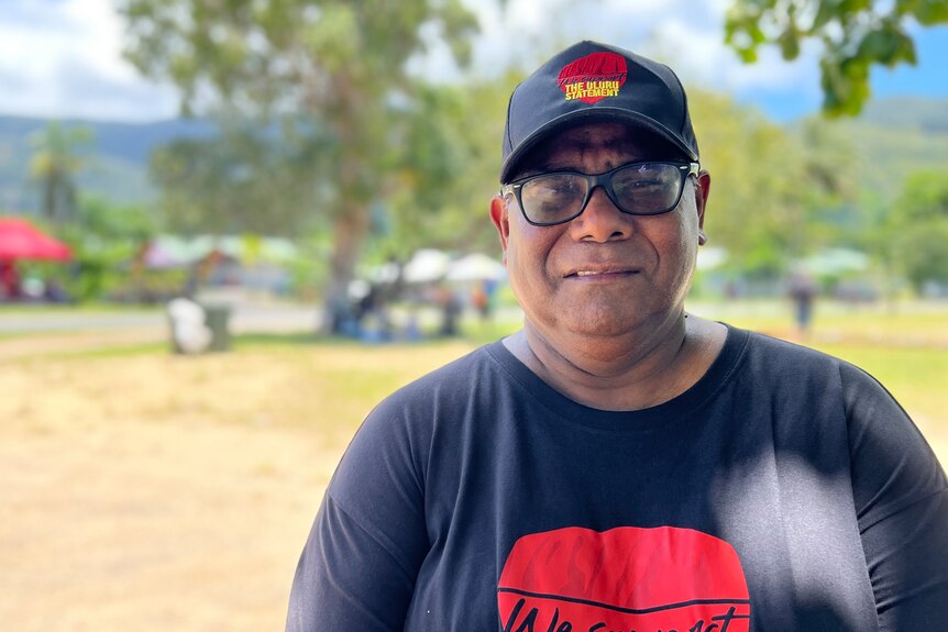 A bespectacled Indigenous man stands outside, wearing a hat and T-shirt in support of the Statement from the Heart.