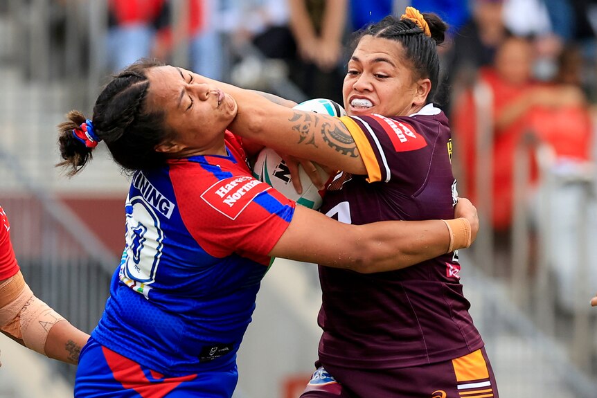 A Brisbane Broncos NRLW player holds the ball while being tackled by a Newcastle Knights opponent.