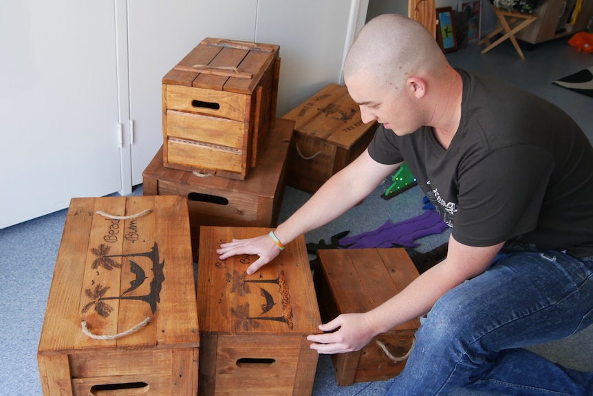 A man kneels on the ground, he is inspecting wooden boxes with artwork on them.
