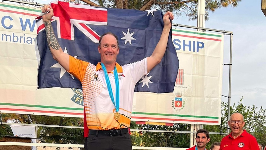 A man in an athletics shirt holds an Australian flag aloft while standing on a podium.